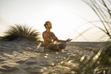 Shirtless young man meditating while practicing yoga on sand at beach against clear sky during sunset - UUF21785