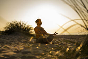 Hemdloser Mann meditiert beim Yoga auf Sand am Strand gegen den klaren Himmel bei Sonnenuntergang - UUF21786