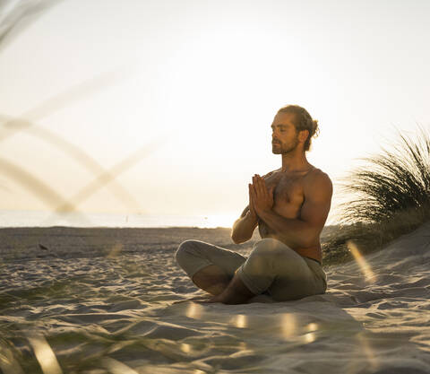 Shirtless young man meditating while practicing yoga sitting with hands clasped at beach against clear sky during sunset stock photo
