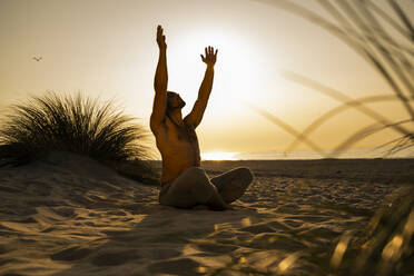 Hemdloser junger Mann übt Yoga, während er mit erhobenen Armen auf dem Sand am Strand gegen den klaren Himmel bei Sonnenuntergang sitzt - UUF21788