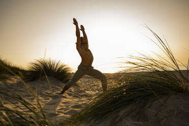Hemdloser junger Mann, der inmitten von Pflanzen am Strand gegen den klaren Himmel bei Sonnenuntergang Yoga in der Kriegerposition praktiziert - UUF21789