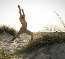 Shirtless junger Mann übt Krieger Position Yoga am Strand gegen klaren Himmel bei Sonnenuntergang - UUF21790