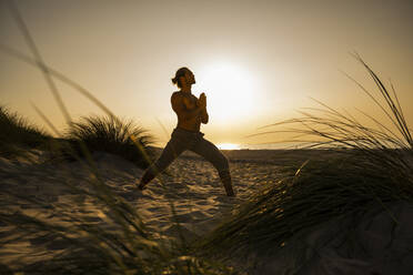 Hemdloser junger Mann übt Yoga in der Kriegerposition mit verschränkten Händen am Strand gegen den klaren Himmel bei Sonnenuntergang - UUF21791