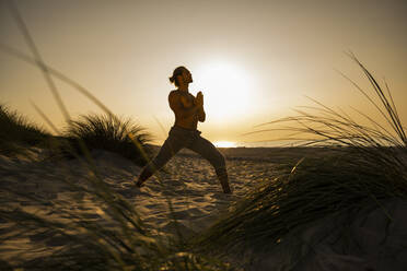 Shirtless young man practicing warrior position yoga with hands clasped at beach against clear sky during sunset - UUF21791