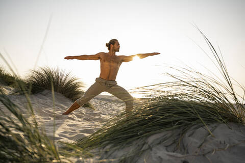 Shirtless young man practicing warrior 2 position yoga at beach against clear sky during sunset stock photo