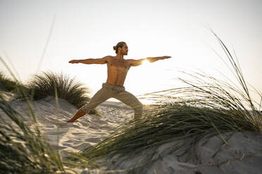 Shirtless junger Mann übt Krieger 2 Position Yoga am Strand gegen klaren Himmel bei Sonnenuntergang - UUF21794