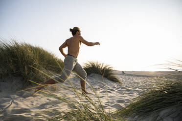 Hemdloser junger Mann läuft am Strand inmitten von Pflanzen gegen den klaren Himmel bei Sonnenuntergang - UUF21798