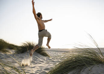 Shirtless young man jumping amidst plants at beach against clear sky during sunset - UUF21799