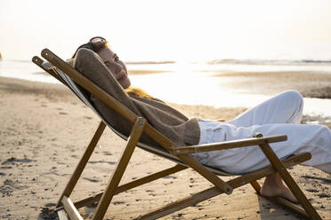 Schöne junge Frau entspannt sich auf einem Klappstuhl am Strand bei Sonnenuntergang - UUF21800