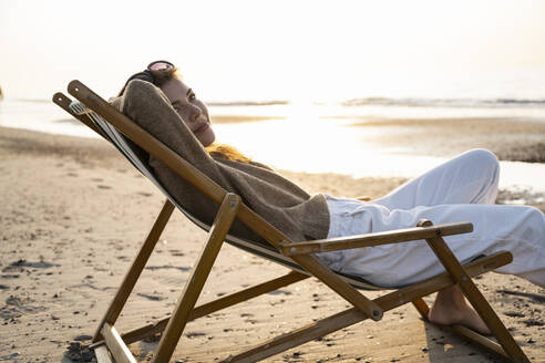 Beautiful young woman relaxing on folding chair at beach during sunset - UUF21800