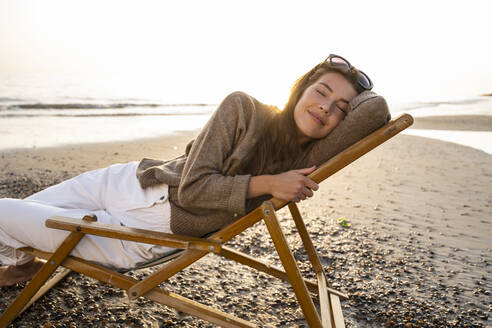 Smiling beautiful young woman reclining on folding chair while relaxing at beach during sunset - UUF21801