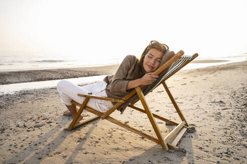 Relaxed woman reclining on folding chair at beach against clear sky during sunset - UUF21802