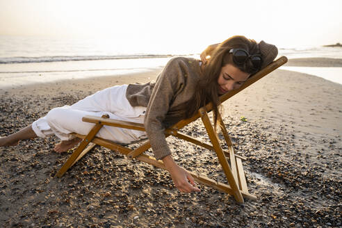 Young woman reclining on folding chair at beach against clear sky during sunset - UUF21803