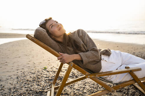 Thoughtful woman reclining on folding chair at beach against clear sky during sunset - UUF21804