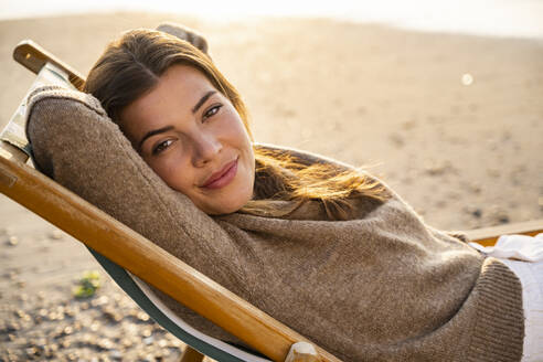 Beautiful young woman reclining on folding chair at beach during sunset - UUF21805