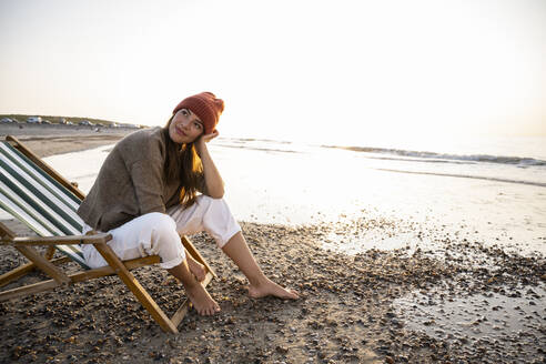 Thoughtful woman sitting on folding chair while looking away at beach against clear sky during sunset - UUF21807