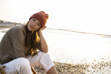 Beautiful young woman wearing knit hat sitting at beach during sunset - UUF21808