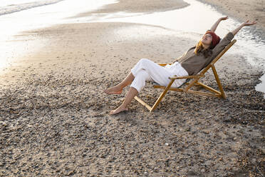 Smiling young woman sitting with legs cross and arms raised on folding chair at beach during sunset - UUF21811