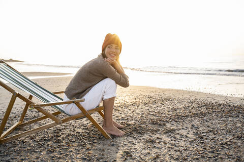 Lächelnde junge Frau, die auf einem Klappstuhl sitzt und bei Sonnenuntergang gegen den klaren Himmel auf den Strand blickt, lizenzfreies Stockfoto