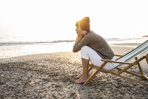 Back lit thoughtful young woman sitting on folding chair while looking away at beach against clear sky during sunset stock photo