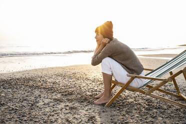 Nachdenkliche junge Frau sitzt auf einem Klappstuhl und blickt bei Sonnenuntergang auf den Strand und den klaren Himmel - UUF21813