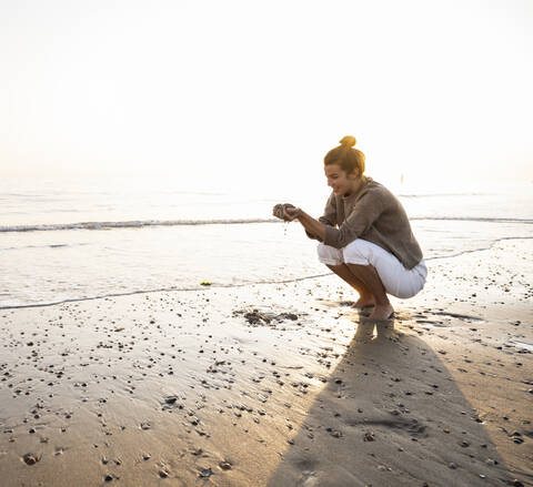 Beautiful young woman crouching while holding sand on shore at beach during sunset stock photo