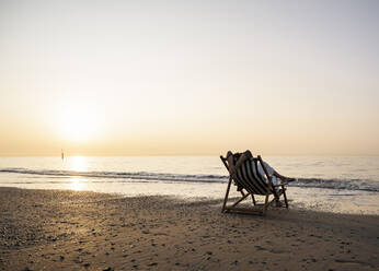 Woman relaxing while sitting on folding chair at beach against clear sky during sunset - UUF21819