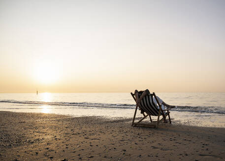 Frau, die sich auf einem Klappstuhl am Strand vor einem klaren Himmel bei Sonnenuntergang entspannt - UUF21819