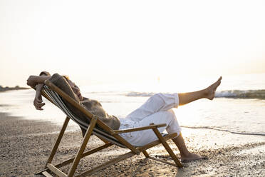 Entspannte junge Frau sitzt auf einem Klappstuhl am Strand bei Sonnenuntergang - UUF21820