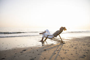 Smiling young woman relaxing while sitting on folding chair at beach against clear sky during sunset - UUF21821