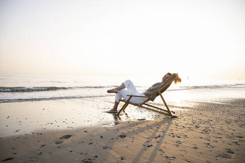 Smiling young woman relaxing while sitting on folding chair at beach against clear sky during sunset - UUF21821