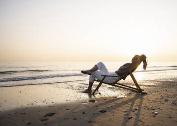 Lächelnde junge Frau entspannt sich auf einem Klappstuhl am Strand gegen den klaren Himmel bei Sonnenuntergang - UUF21822