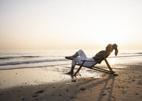 Smiling young woman relaxing on folding chair at beach against clear sky during sunset - UUF21822