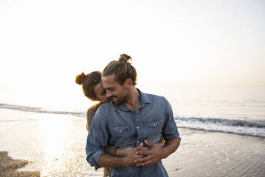 Woman embracing boyfriend from behind at beach against clear sky during sunset - UUF21823