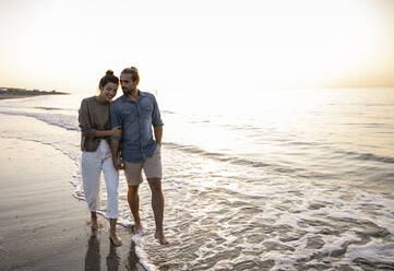 Young couple talking while walking on shore at beach against clear sky during sunset - UUF21825