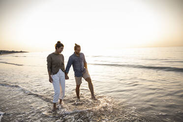 Happy young couple holding hands while walking on shore at beach during sunset - UUF21826