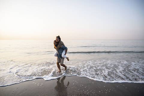 Cheerful man giving piggyback to girlfriend while walking on shore at beach against clear sky during sunset stock photo
