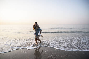 Cheerful man giving piggyback to girlfriend while walking on shore at beach against clear sky during sunset - UUF21829