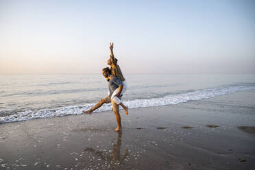 Junger Mann nimmt seine Freundin am Strand gegen den klaren Himmel bei Sonnenuntergang huckepack - UUF21830