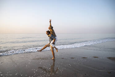 Young man giving piggyback to girlfriend on shore at beach against clear sky during sunset - UUF21830