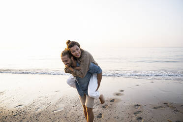 Happy man giving piggyback to girlfriend while walking on shore at beach against clear sky during sunset - UUF21831