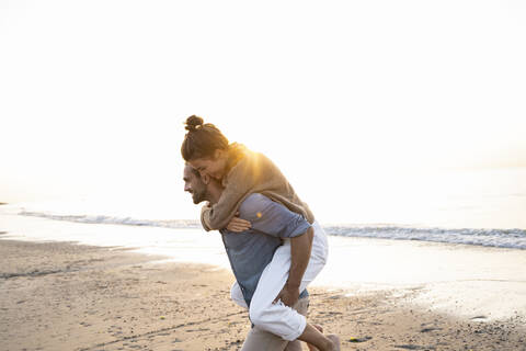 Smiling man giving piggyback to girlfriend while walking on shore at beach against clear sky during sunset stock photo