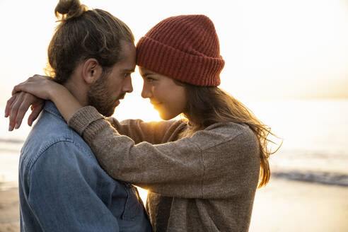Verliebtes junges Paar, das bei Sonnenuntergang von Angesicht zu Angesicht am Strand steht - UUF21838