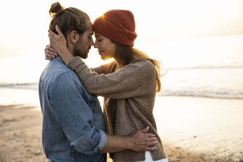 Junge Frau, die ihren Freund umarmt, während sie bei Sonnenuntergang am Strand steht - UUF21839