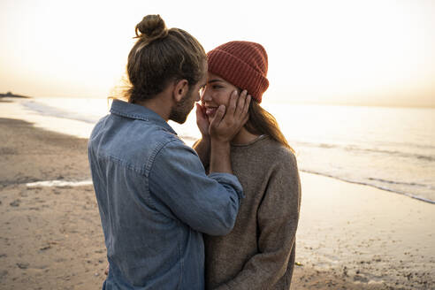 Romantischer junger Mann, der das Gesicht seiner Freundin berührt, während er bei Sonnenuntergang am Strand steht - UUF21841