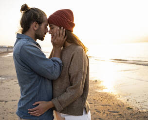 Romantic young man standing face to face with girlfriend at beach during sunset - UUF21842