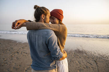 Junges romantisches Paar steht von Angesicht zu Angesicht am Strand gegen den klaren Himmel bei Sonnenuntergang - UUF21844