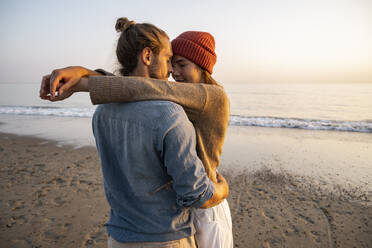 Young romantic couple standing face to face at beach against clear sky during sunset - UUF21844