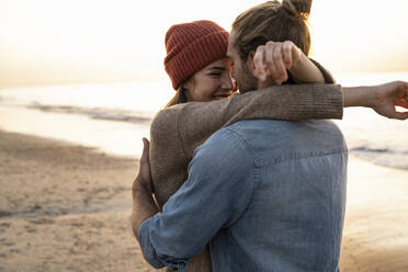 Smiling young woman embracing boyfriend while standing at beach during sunset - UUF21845