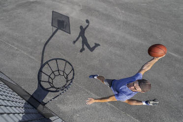 Junger Mann, der den Ball in den Reifen eintaucht, während er an einem sonnigen Tag Basketball spielt - STSF02620